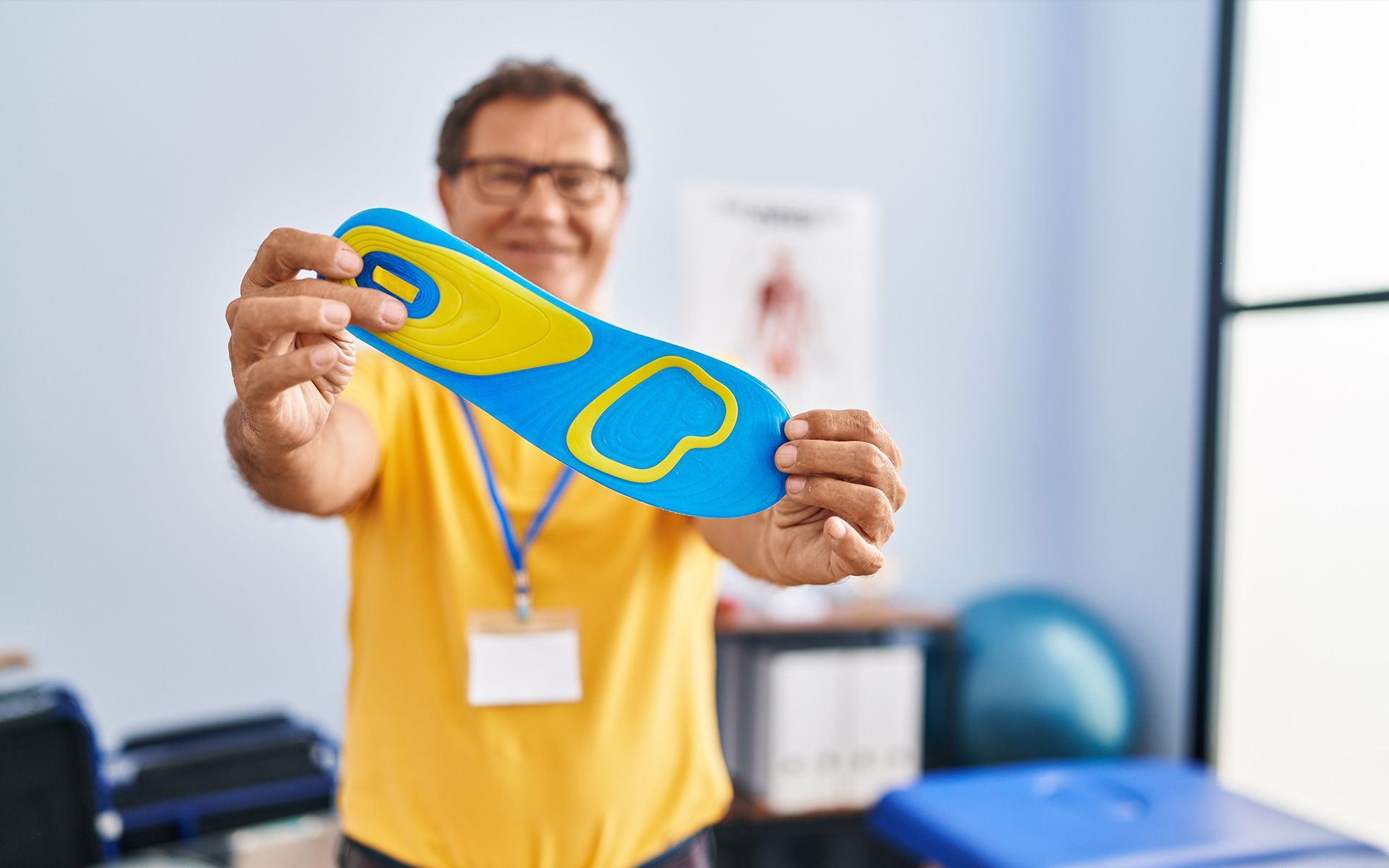 Man in yellow shirt holding blue and yellow orthotic in clinic.