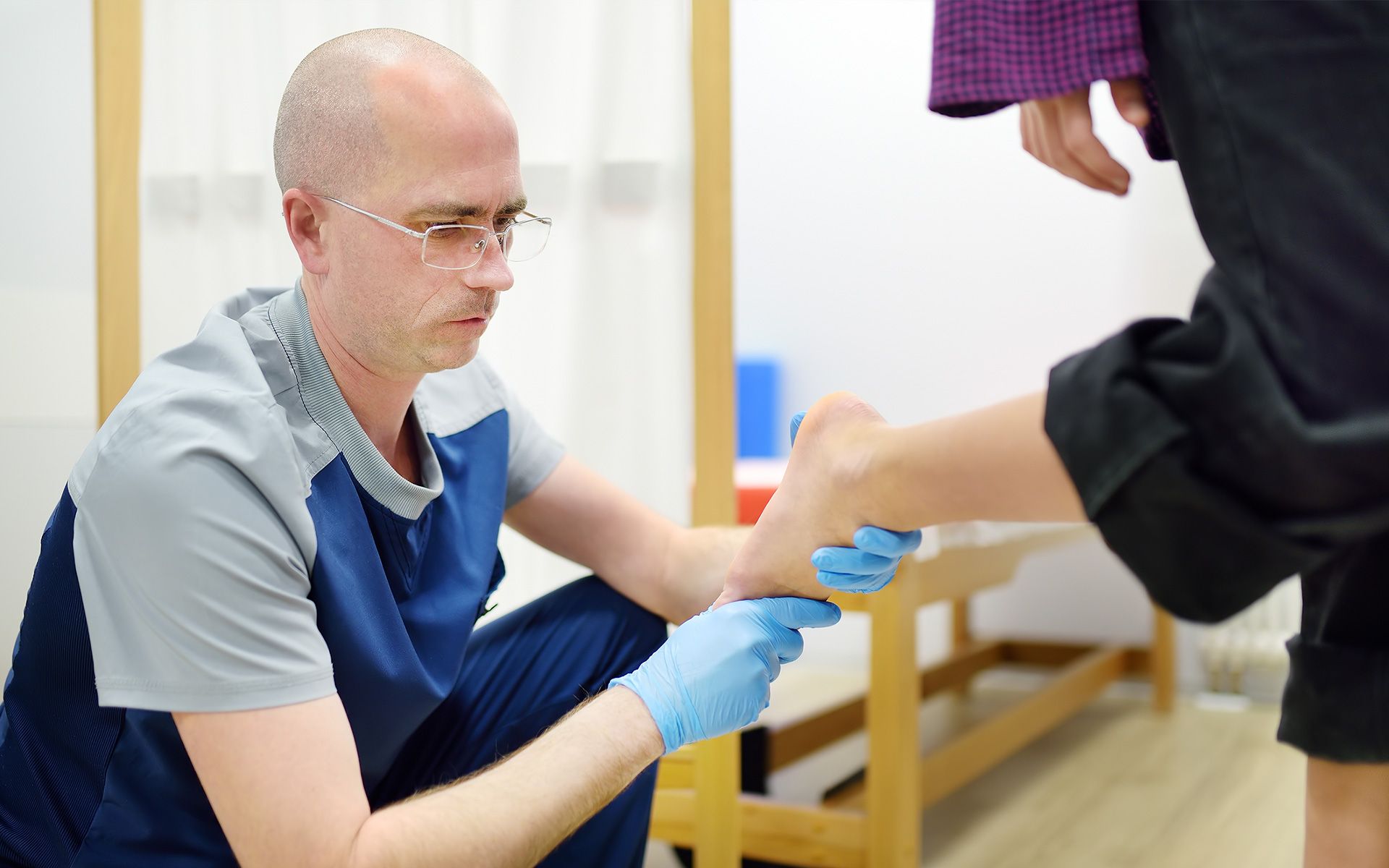 Podiatrist examining a patient's foot; indoors. Doctor wears gloves, patient in dark pants.