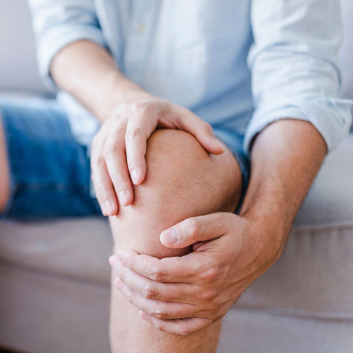 Man seated, holding knee with hands, possibly in pain, indoors.