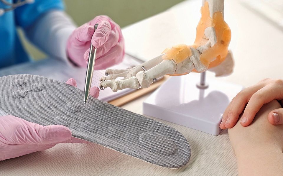 A medical professional examining a gray orthotic insole, pointing with tweezers, near a foot skeleton model.