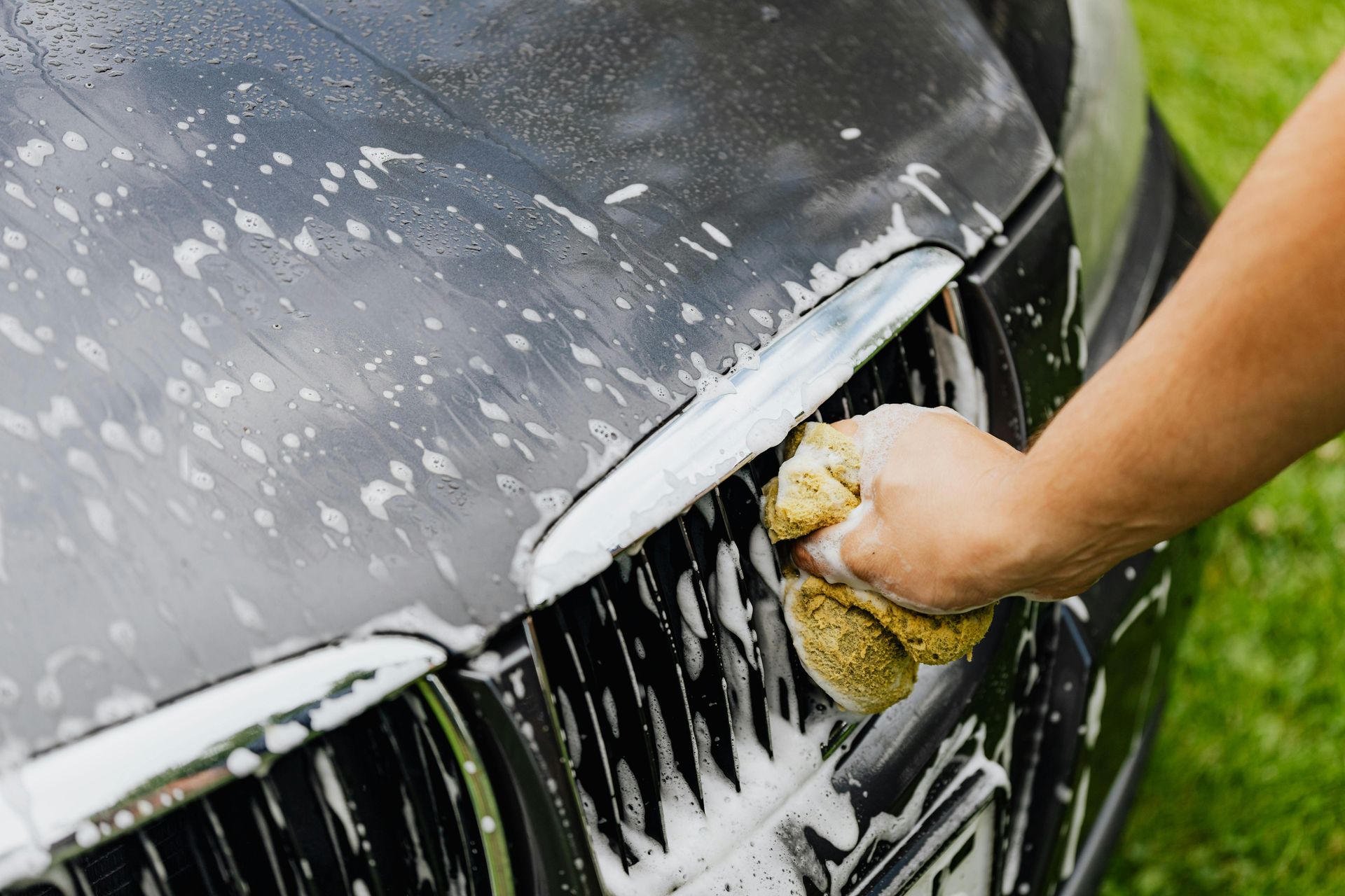 Hand washing a black car's grill with a soapy sponge outdoors on a grassy lawn.