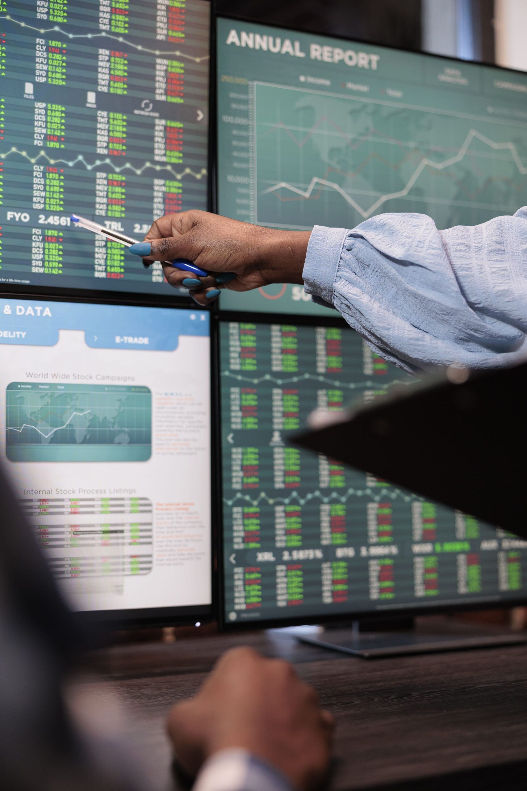 Person pointing at stock market data on multiple computer screens.