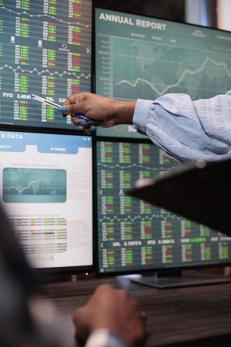 Person pointing at stock market data on multiple computer screens.