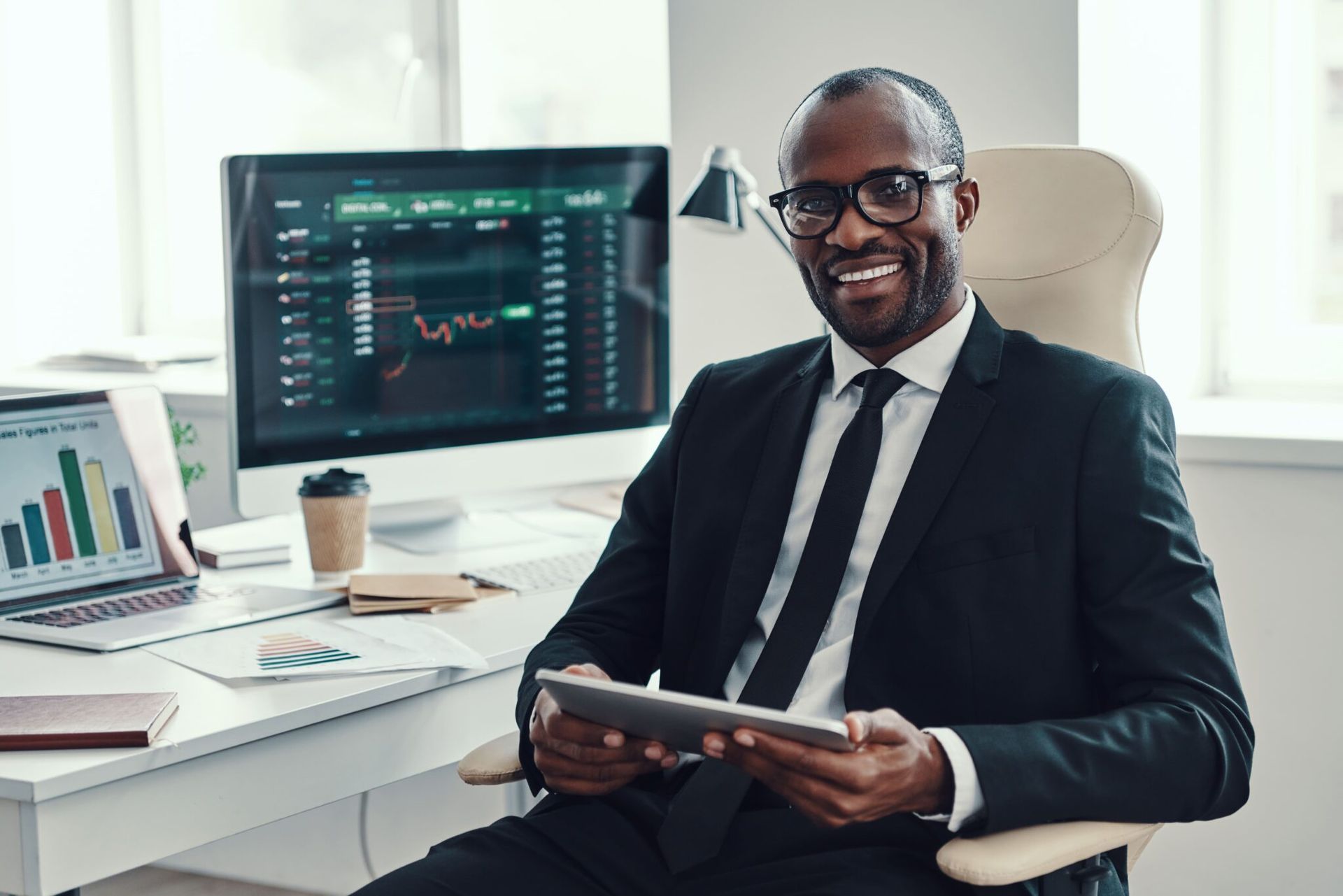 Smiling Black businessman in suit at desk with computer and tablet.