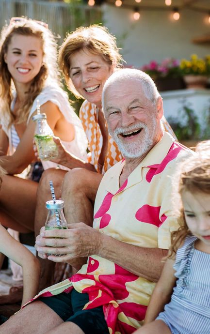 Family smiling together outdoors. Older man in floral shirt holds drink, other family members visible.
