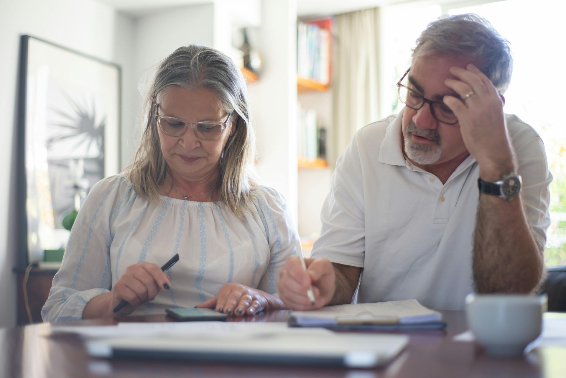 Older couple reviews financial documents, looking concerned, at a table indoors.