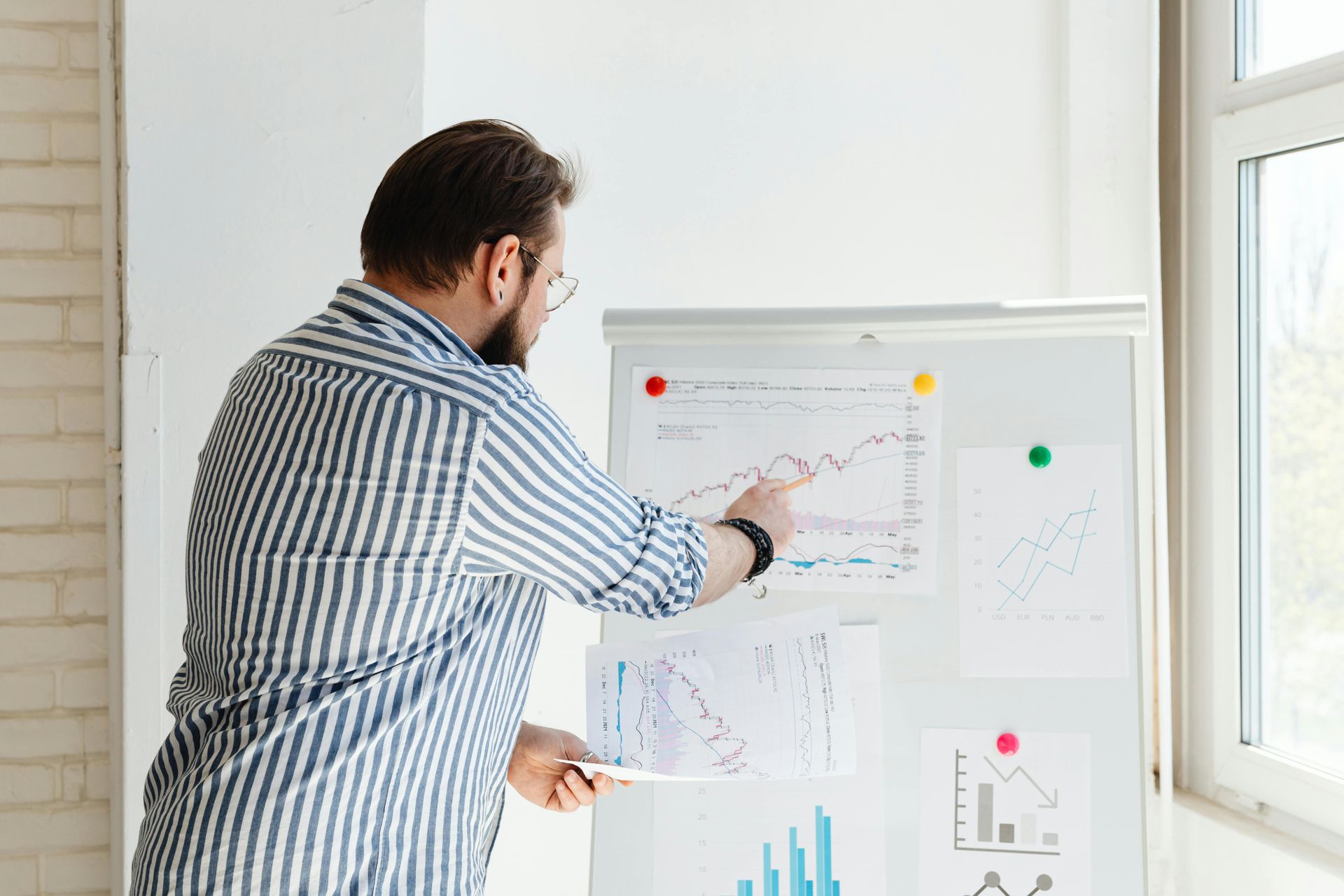 Man in striped shirt points to a graph on a whiteboard, presenting data in a bright office.