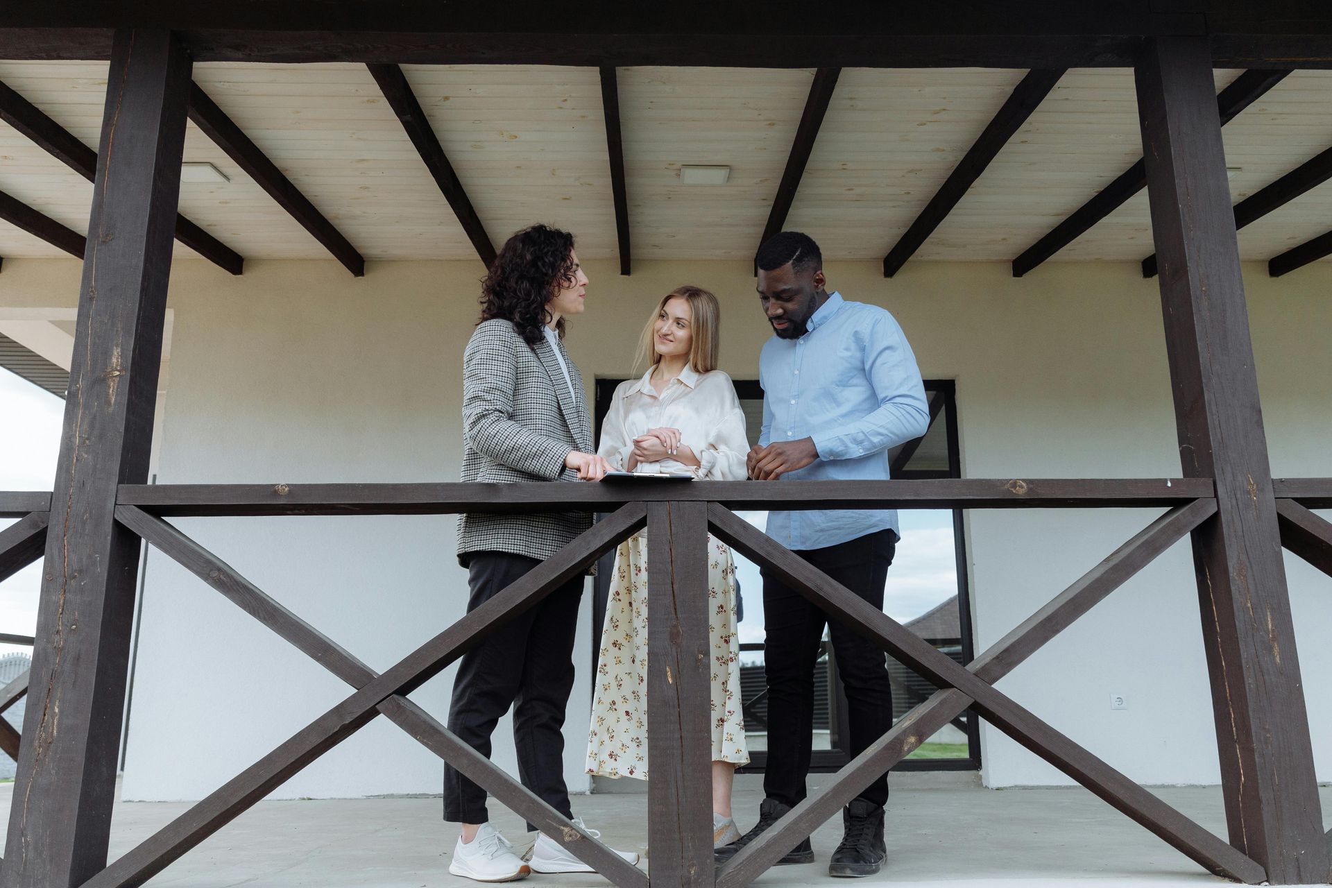 Three people on a porch, conversing. A woman in the middle, flanked by a man on each side, looking at papers.