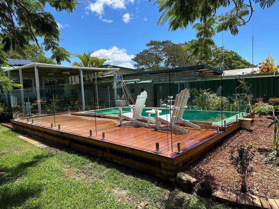 A swimming pool with a wooden deck and chairs around it. — Glass Pool Fencing Australia in Wauchope, NSW