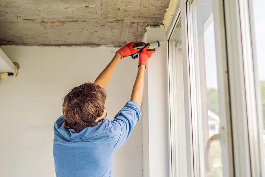 A person wearing red gloves uses a caulk gun to seal the top edge of a window frame in an unfinished room.