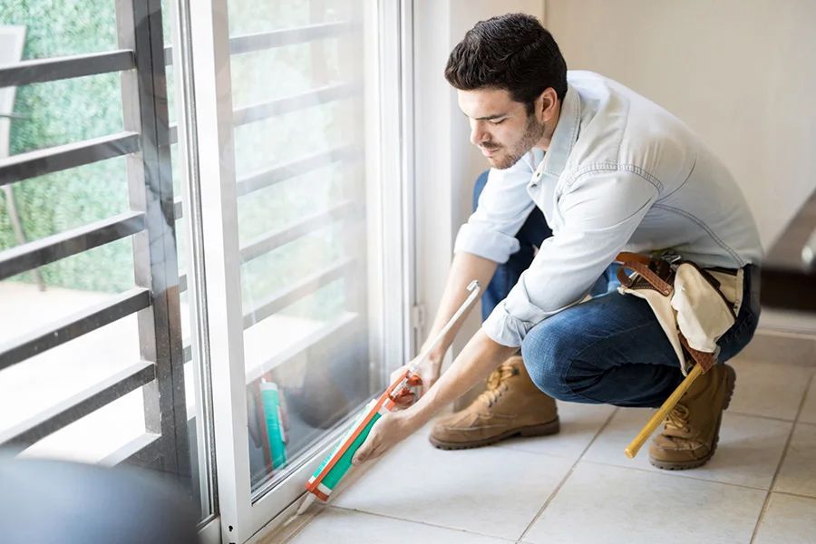 A person in work clothes kneeling to apply caulk to the base of a glass sliding door frame.