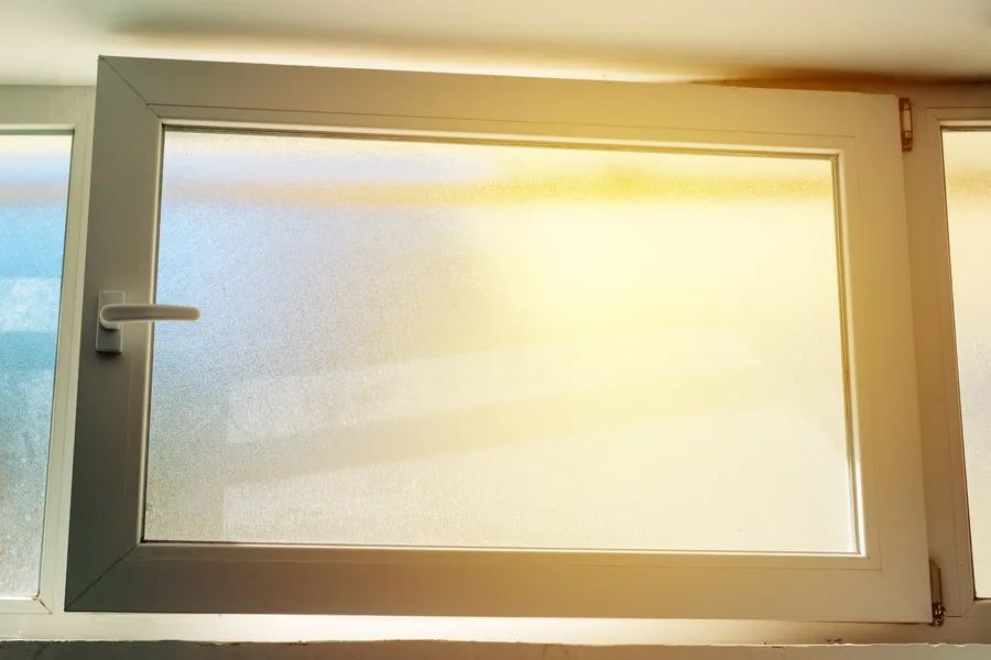 A white-framed window with frosted glass is slightly ajar, with bright, warm sunlight glowing through the pane.