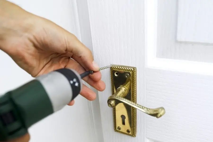 A person uses a handheld power drill to attach a brass-colored door handle to a white door.