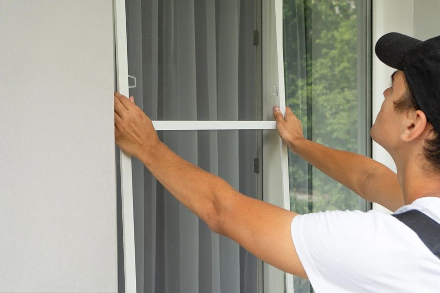A person in a cap installs a white framed screen onto a glass door or window.