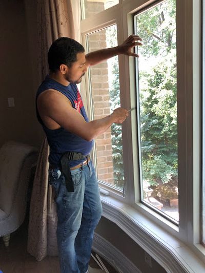 A person in a blue tank top and jeans installs a window pane in a frame, working indoors near curtains and a brick wall.