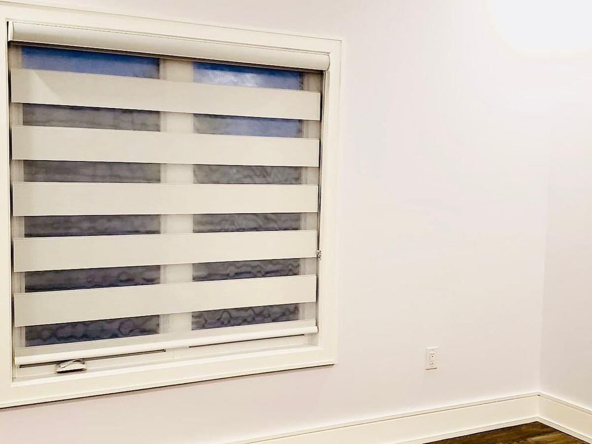 A window featuring white horizontal zebra-style roller shades against a plain white wall with baseboard molding.