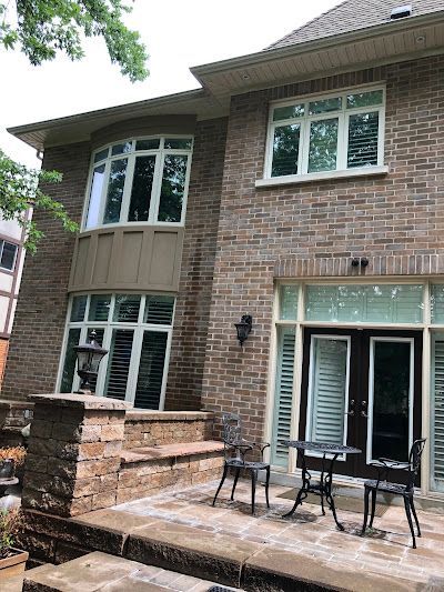 A brick house exterior with a patio, two chairs, a small table, and a built-in stone bench under large windows.
