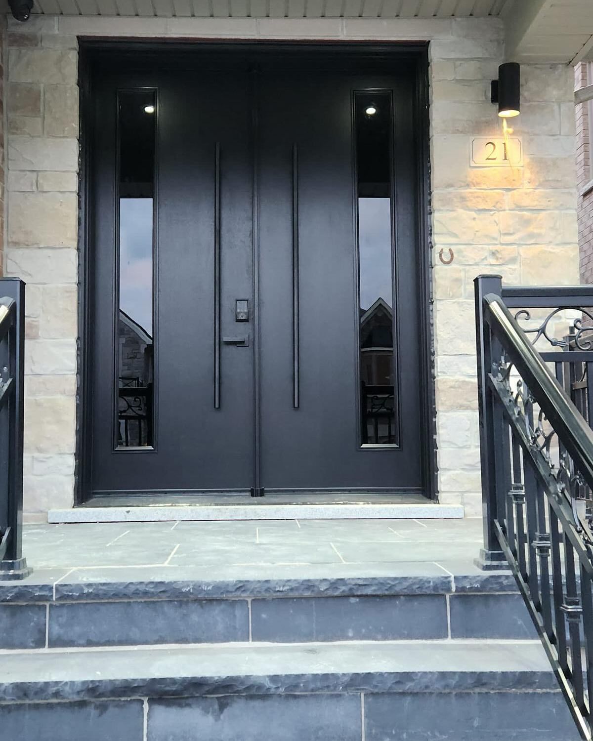Modern black double front doors with vertical glass panels, flanked by stone walls and stone steps at a home entrance.