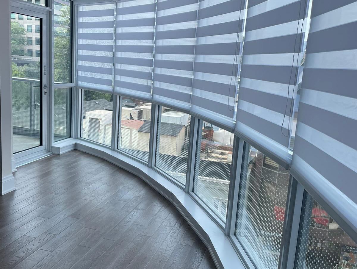 Curved floor-to-ceiling windows with modern gray and white striped roller shades in a room with hardwood floors.