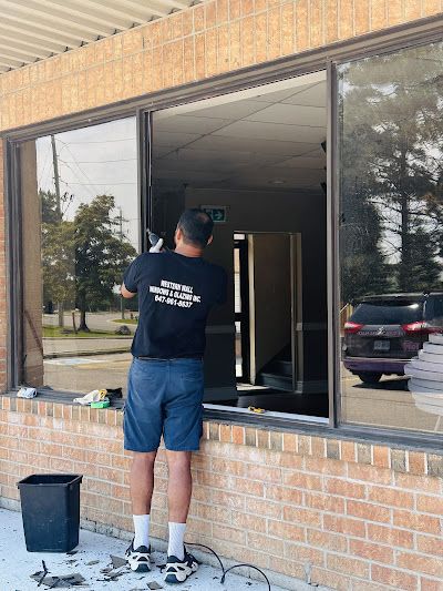 A person wearing a black shirt and blue shorts stands outside a brick building, removing a glass window pane.