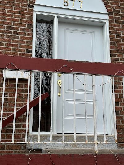 A brick exterior with a white front door labeled 877, framed by a window and a worn white metal railing in the foreground.