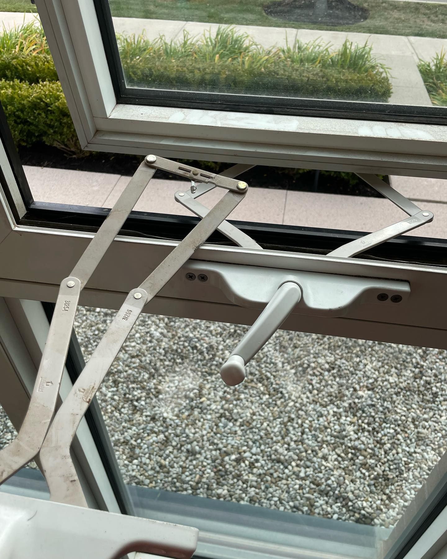 A white crank handle and metal hinge mechanism attached to a beige window frame that is partially open.