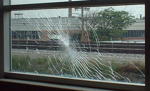 A windowpane with a dense, spiderweb-like crack pattern centered on the glass, looking out onto train tracks and a building.