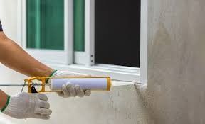 A pair of gloved hands uses a caulking gun to seal the gap between a window frame and a concrete wall.