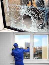 A shattered windowpane next to a worker in blue uniform repairing an intact window.