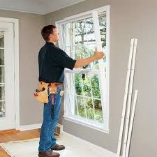 A person in work clothes installs a window trim component onto the side of a white-framed window in a room.