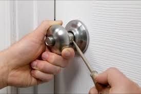 Hands holding a screwdriver to release a metal doorknob from a white door.