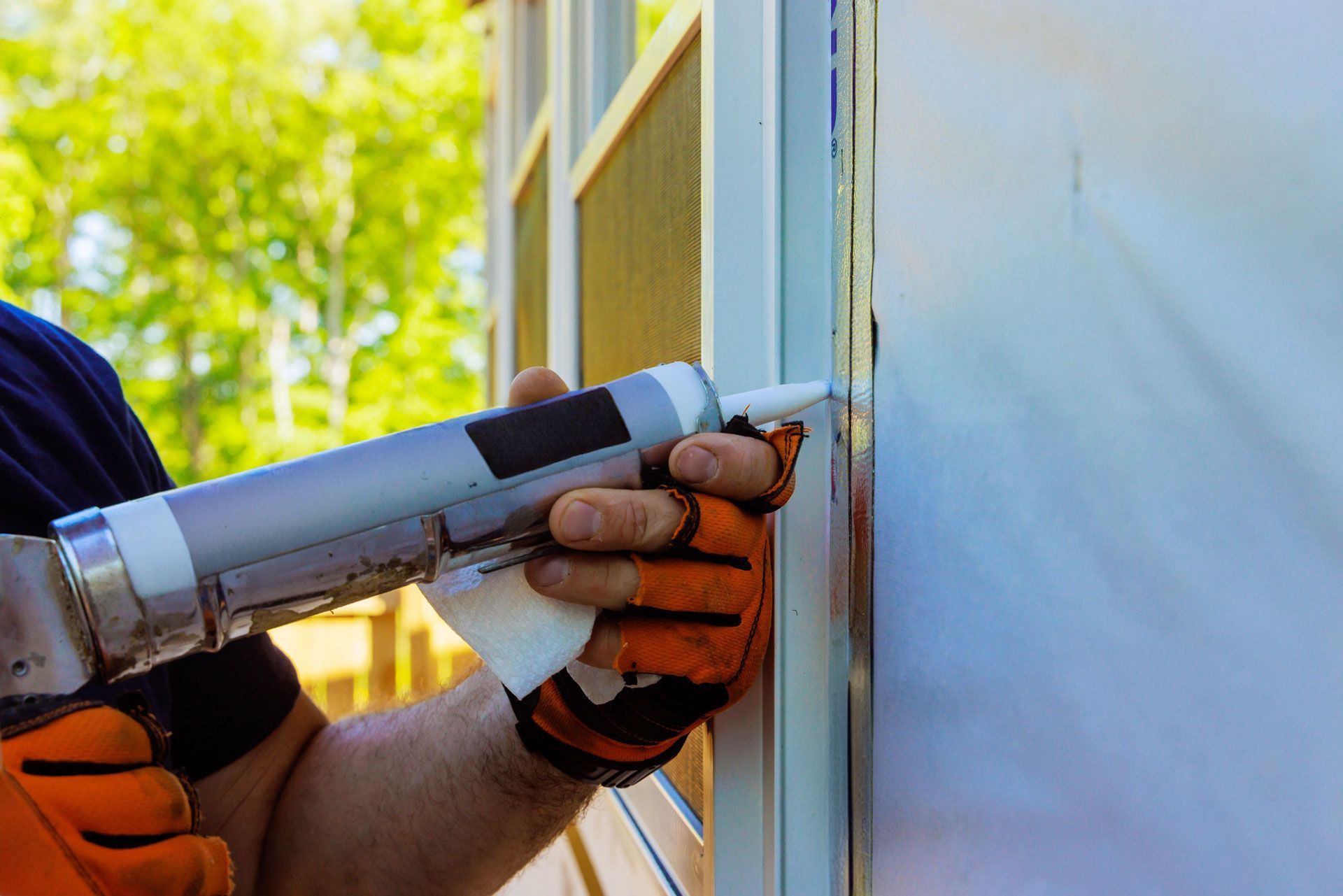 A person wearing orange work gloves uses a caulk gun to apply sealant along the frame of a window or door.
