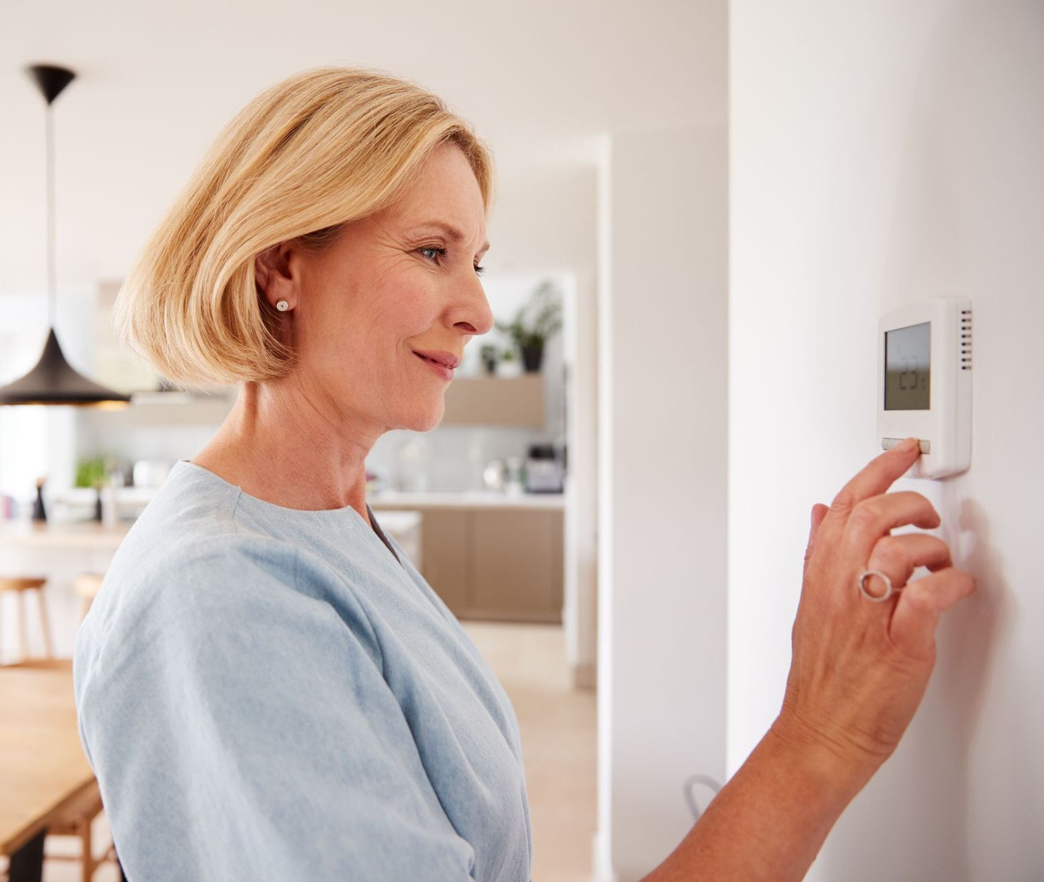 Dans une cuisine bien éclairée, une personne vêtue d'un haut bleu clair règle un thermostat mural.