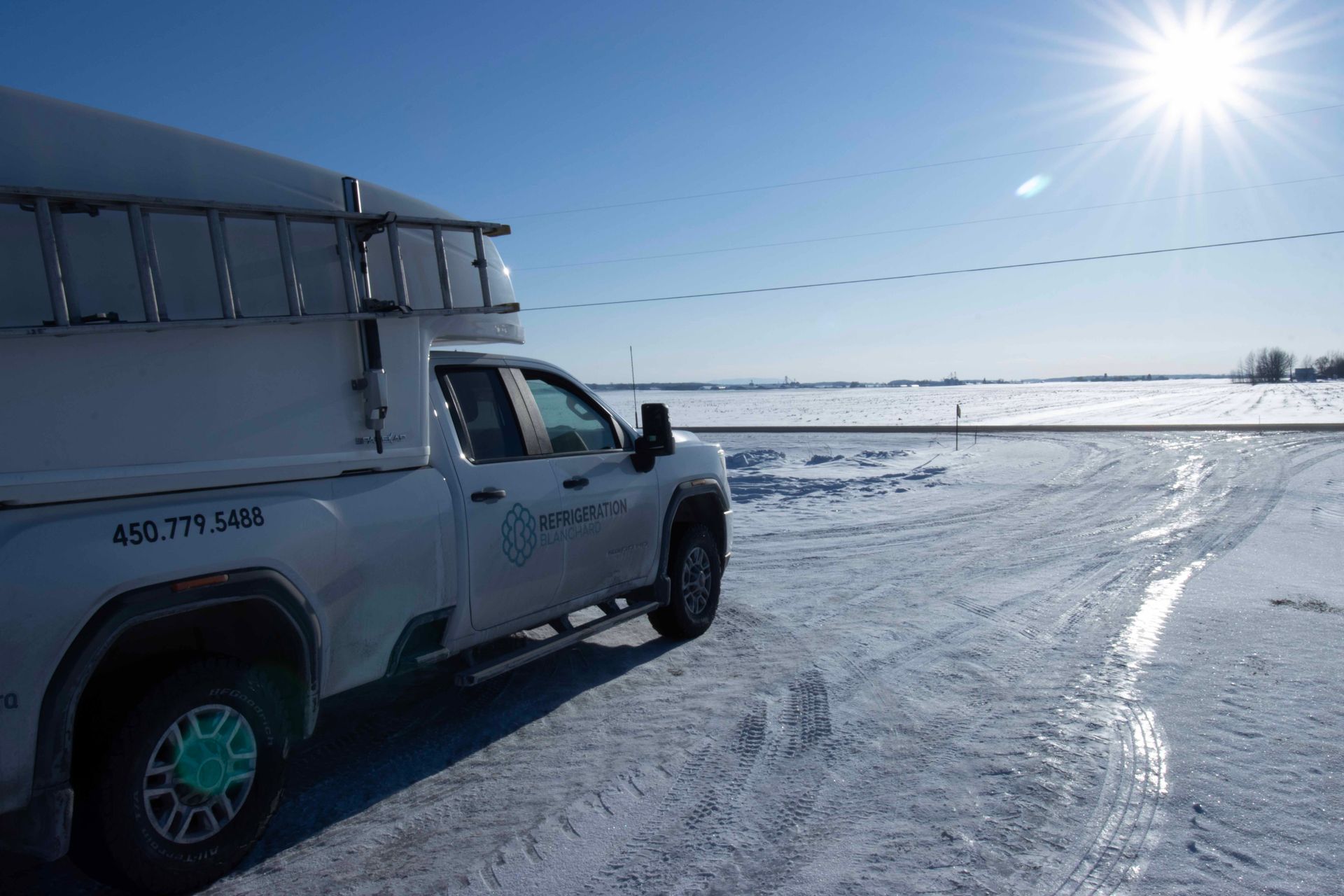 Un camion de dépannage blanc avec une échelle est stationné dans un champ enneigé sous un soleil éclatant.