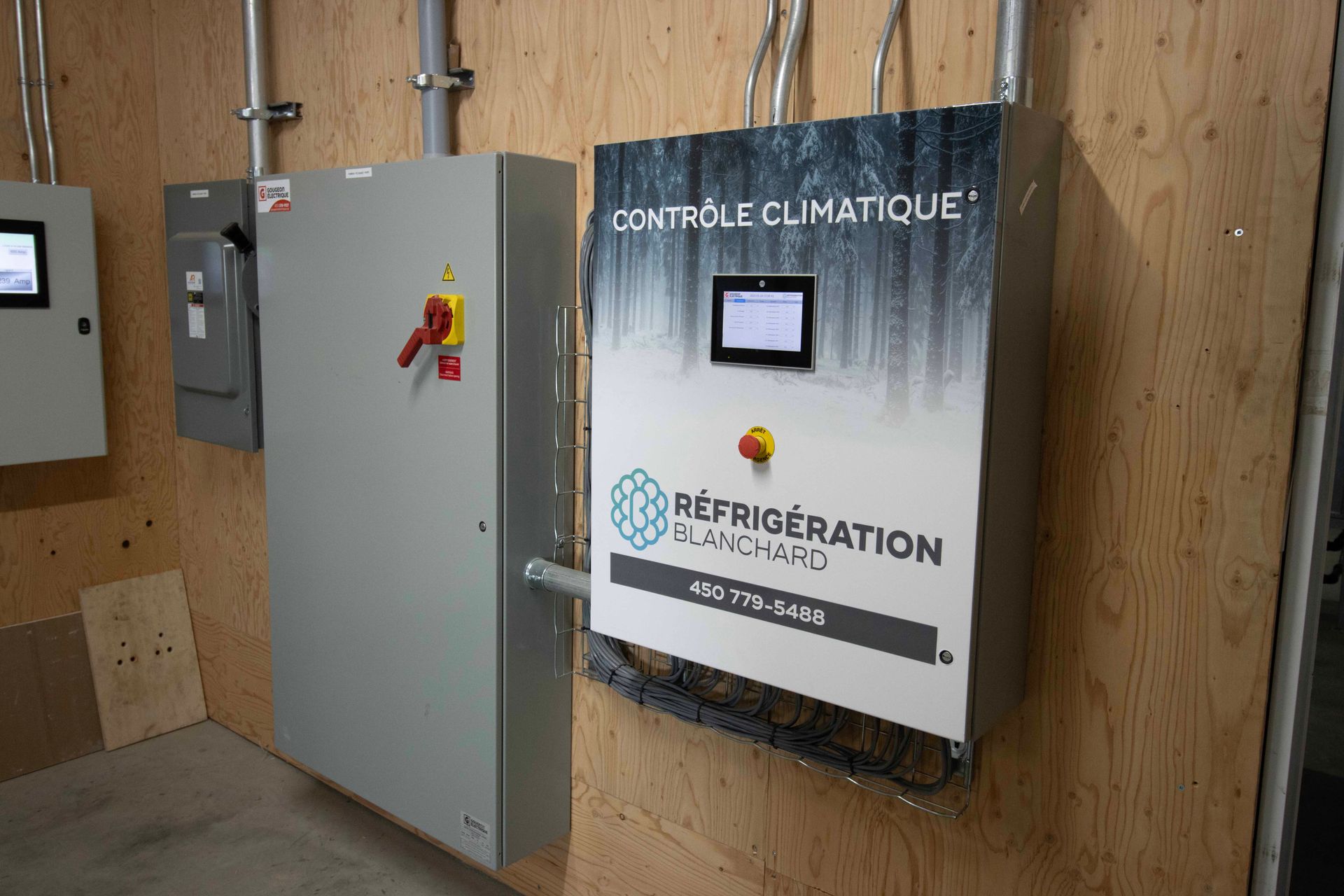 Un panneau de commande climatique de Réfrigération Blanchard monté sur un mur en contreplaqué à côté d'un boîtier de déconnexion électrique.