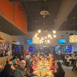Busy restaurant dining room with people seated at long tables under warm pendant lights and industrial ceiling ducts