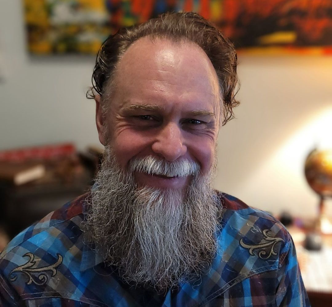 A man with a beard and mustache is smiling and wearing a plaid shirt.