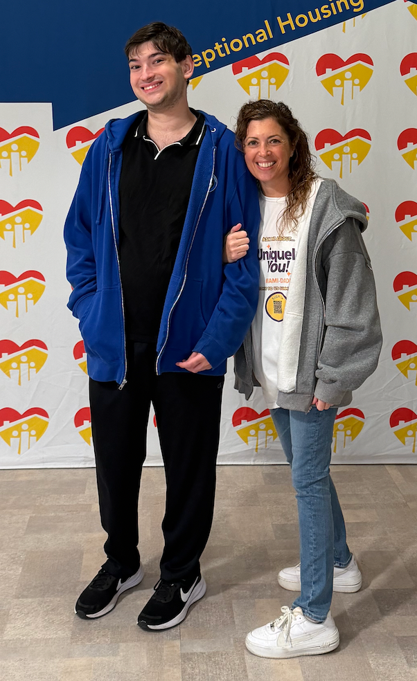 Two people stand side-by-side smiling in front of a backdrop featuring a repeating heart-shaped logo.