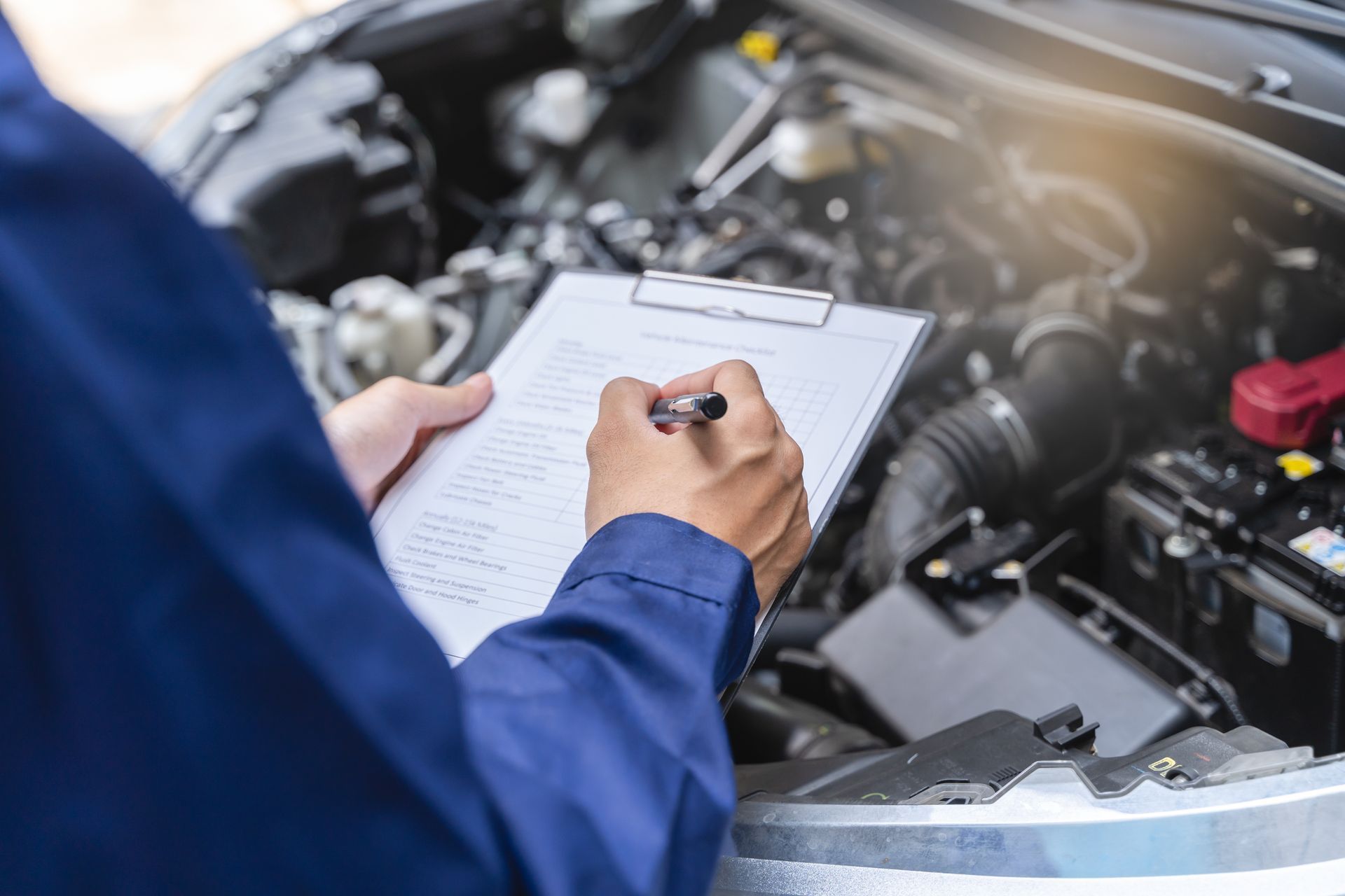Ein Mechaniker in blauem Overall macht sich Notizen, während er einen Automotor unter der Motorhaube untersucht.