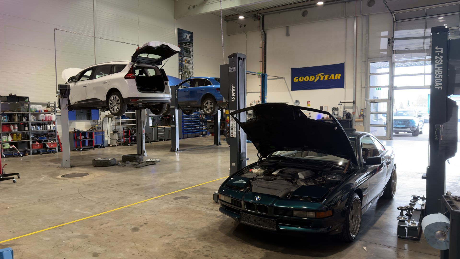 Autowerkstatt mit mehreren Autos auf Hebebühnen und einem dunkelgrünen Auto mit geöffneter Motorhaube im Vordergrund.
