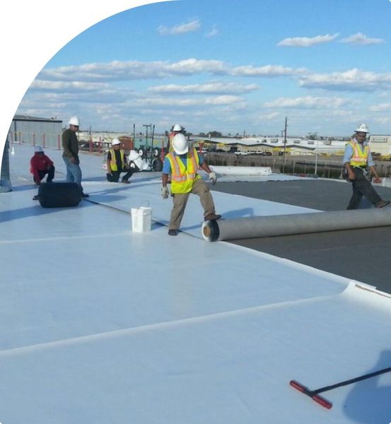 Construction workers installing roofing on a sunny day; they are using a large roll of material, wearing safety gear.