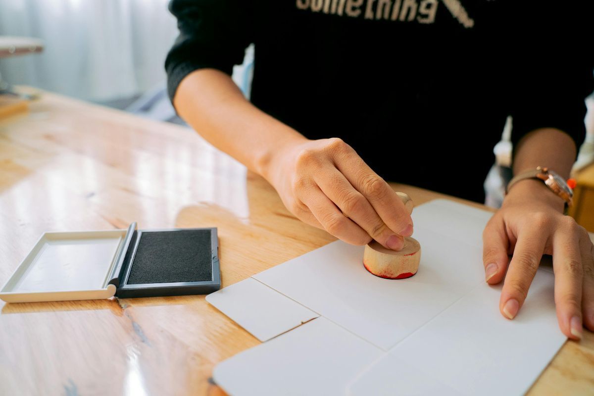 a person’s hand pressing a wooden stamp onto a white paper document on a wooden desk, with a red ink pad partially visible