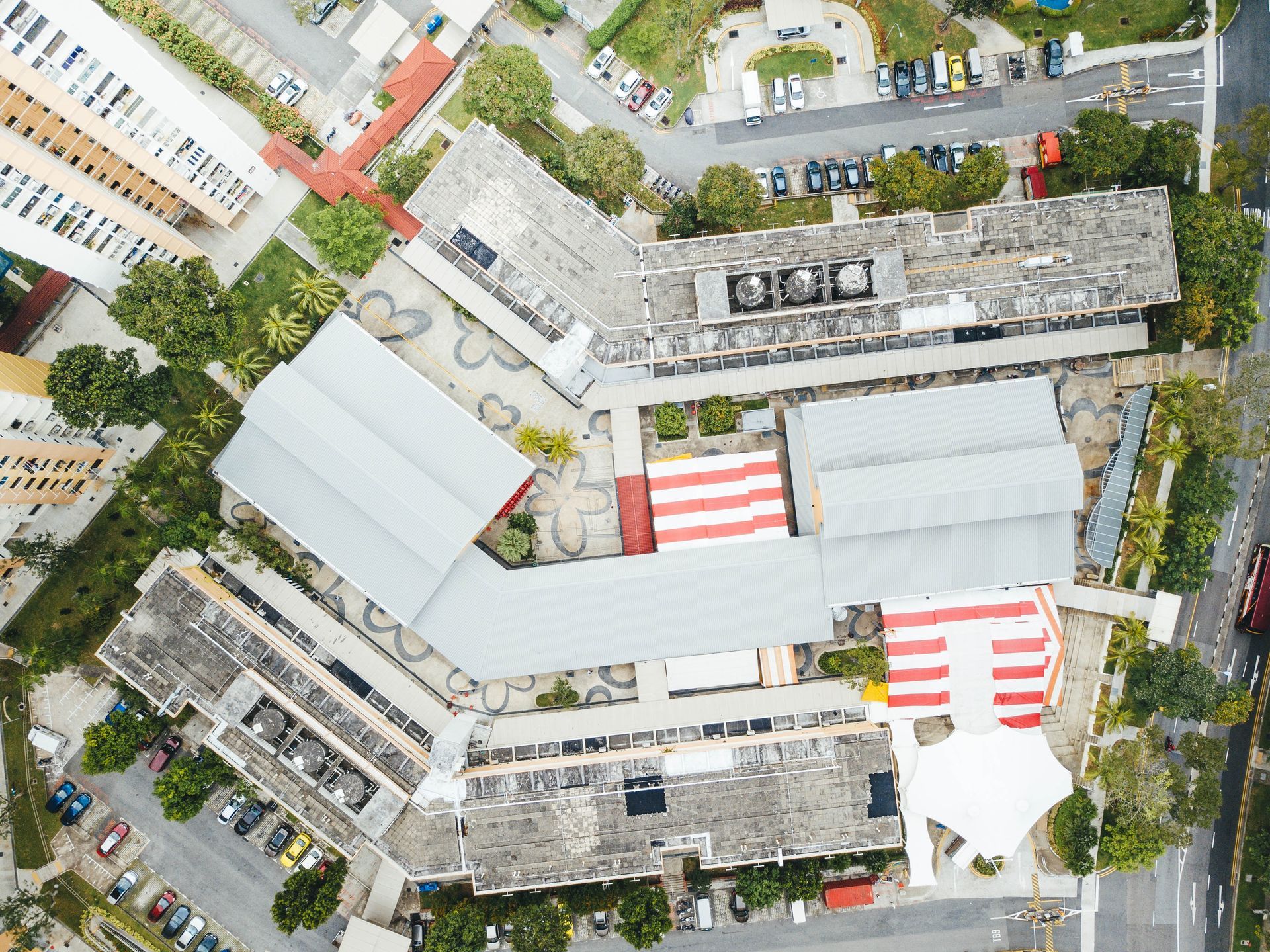 Aerial view of a school complex with gray rooftops, surrounded by trees and parked cars.