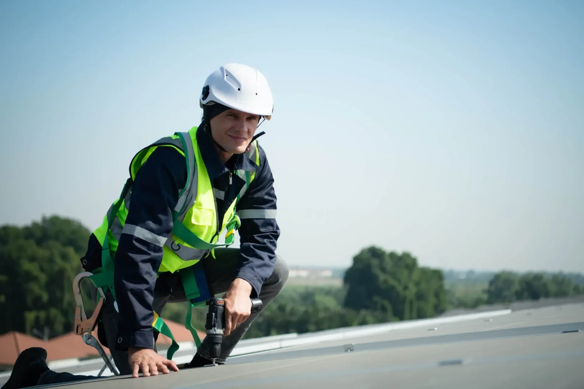 Construction worker in safety gear crouches on a rooftop, smiling at the camera.