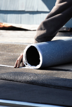 Person unrolling a roll of dark-colored roofing material on a flat roof; hand visible.