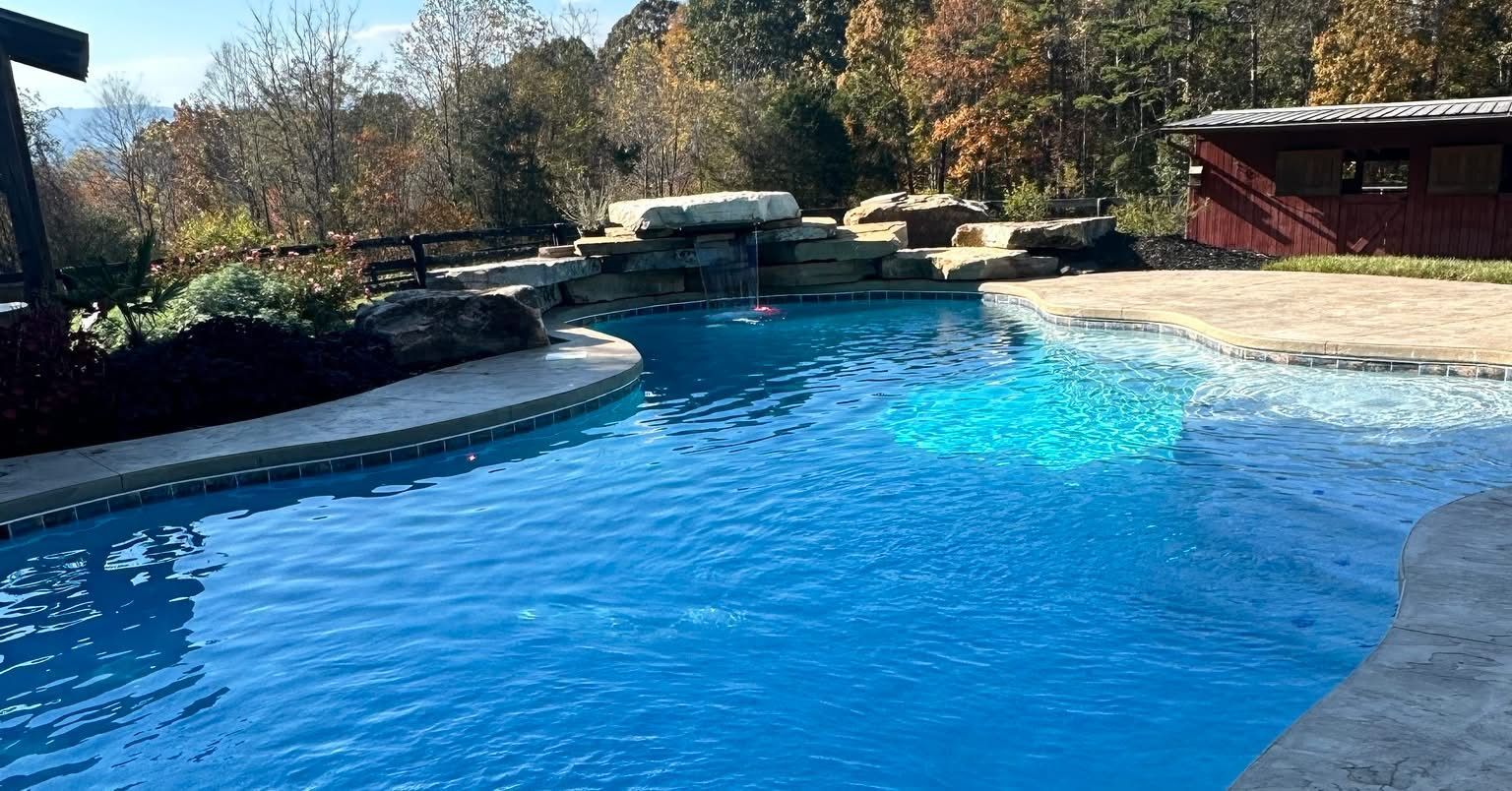 Blue swimming pool with a stone waterfall feature, surrounded by landscaping, trees, and a wooden structure.