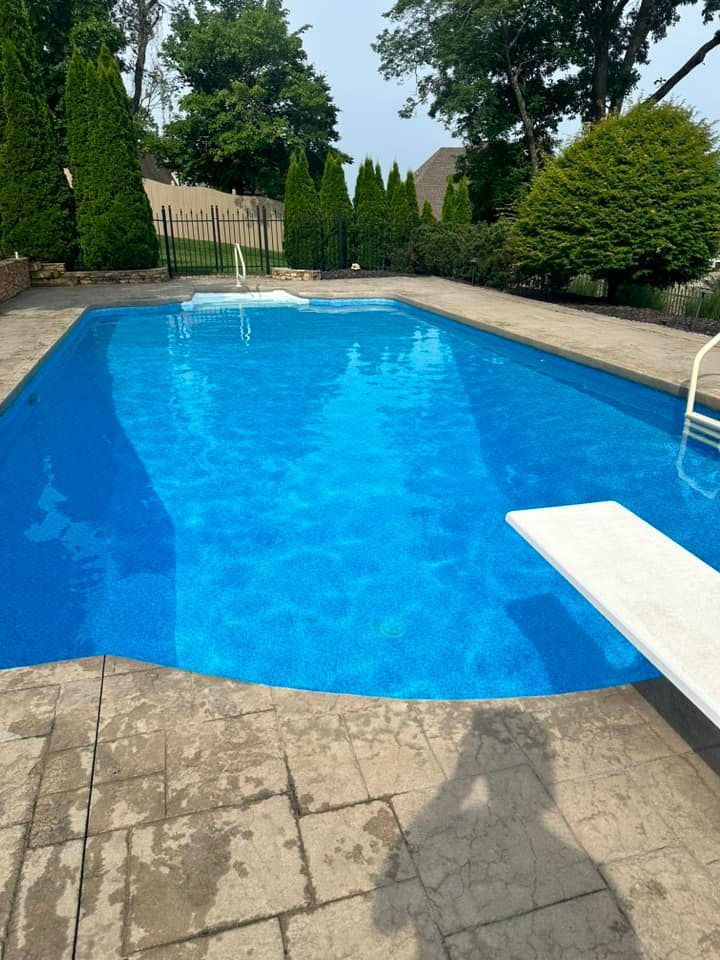 Blue swimming pool with a diving board, surrounded by stone patio and greenery.