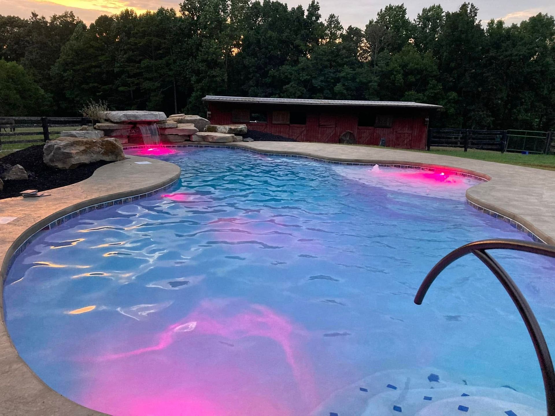 Swimming pool with waterfall, red and blue lights, surrounded by trees and a building.