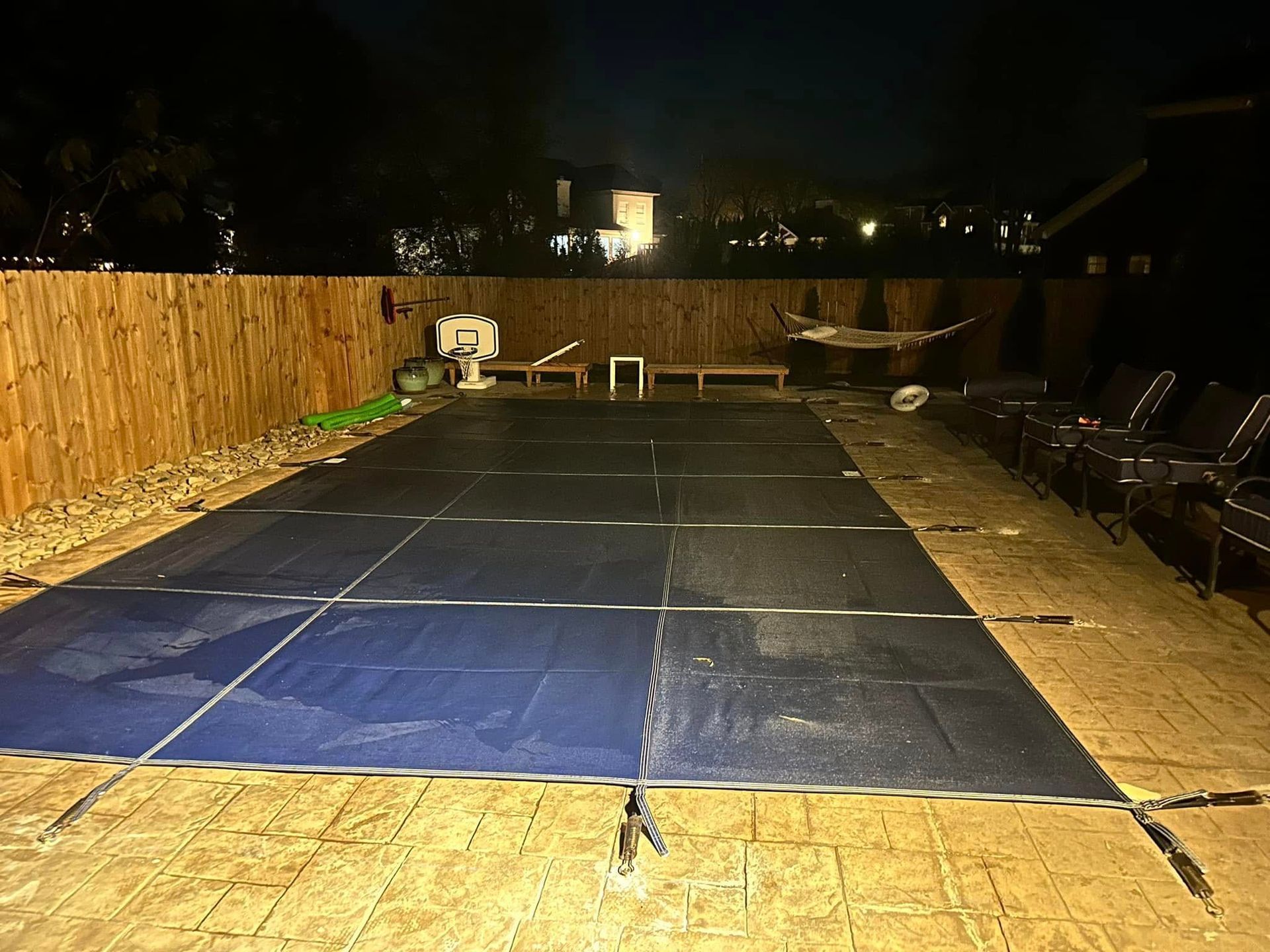 Nighttime backyard scene: a pool covered by a blue tarp, wooden fence, patio furniture, and hammock in the background.