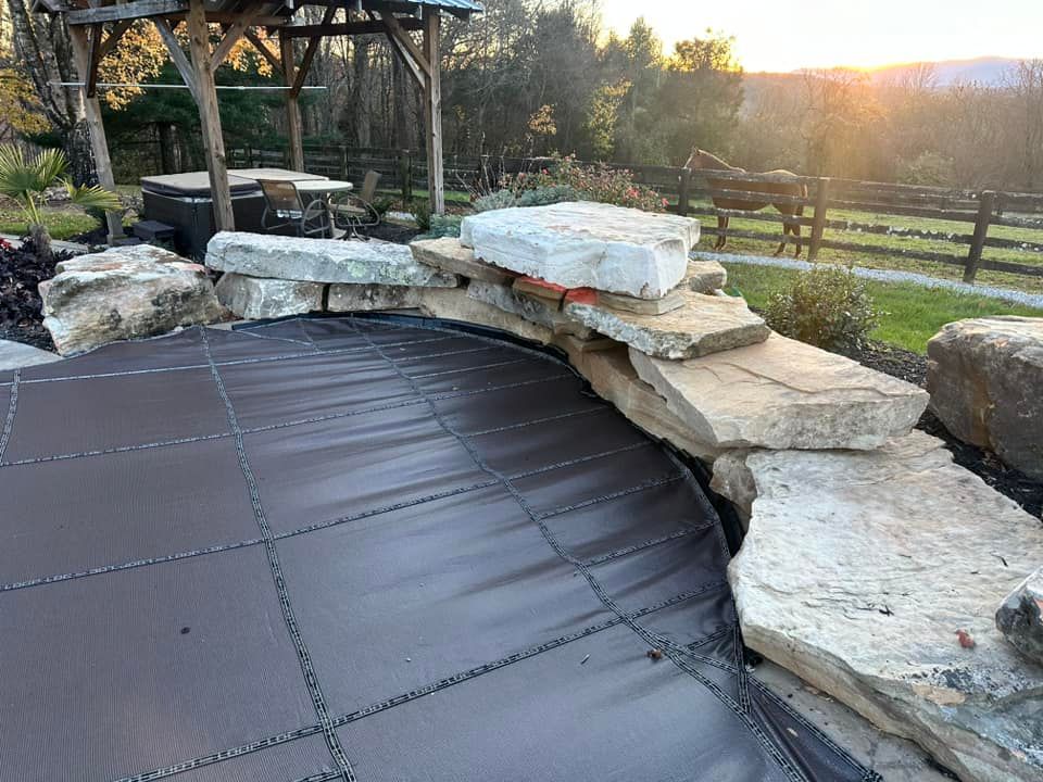 Pool covered with a brown cover, surrounded by stone wall and rocks, with a wooden structure overhead.